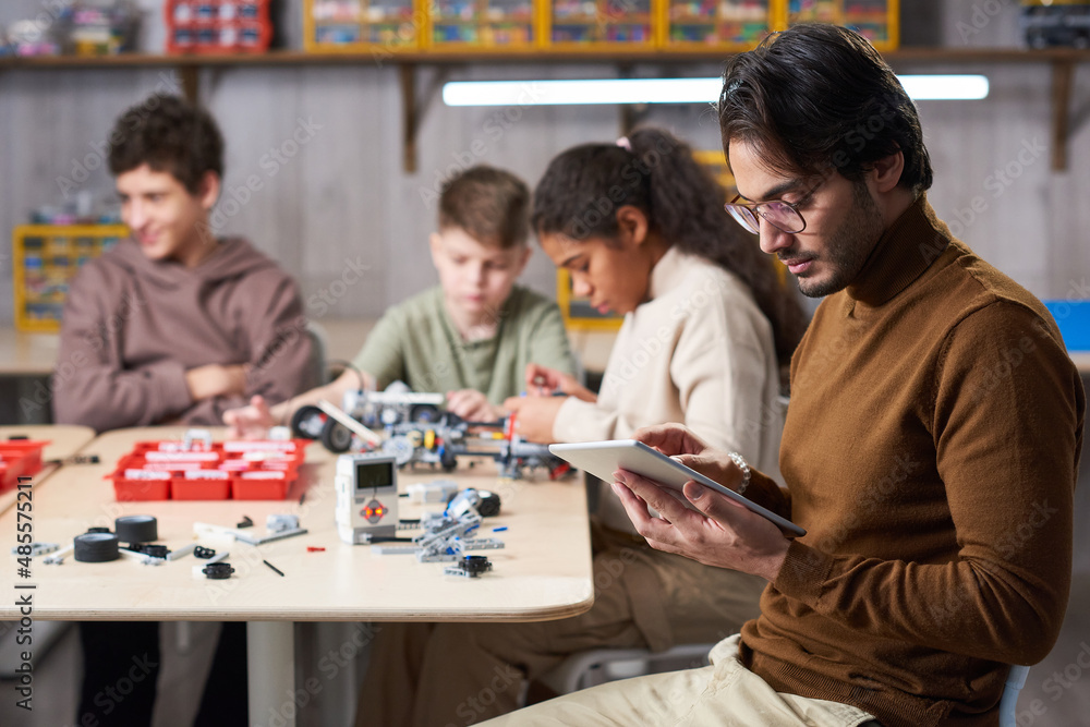 Side view portrait of young male teacher using digital tablet with group of children building robots in background at engineering class, copy space Side view portrait of young male teacher using digital tablet with group of children building robots in background at engineering class, copy space
