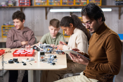 Side view portrait of young male teacher using digital tablet with group of children building robots in background at engineering class  copy space