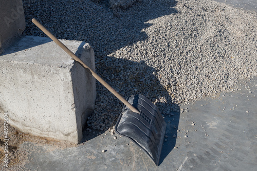 A shovel, leaning against a concrete block next to a huge pile of gray gravel, and the tracks of a truck's tires are visible on the cement floor screed next to it.