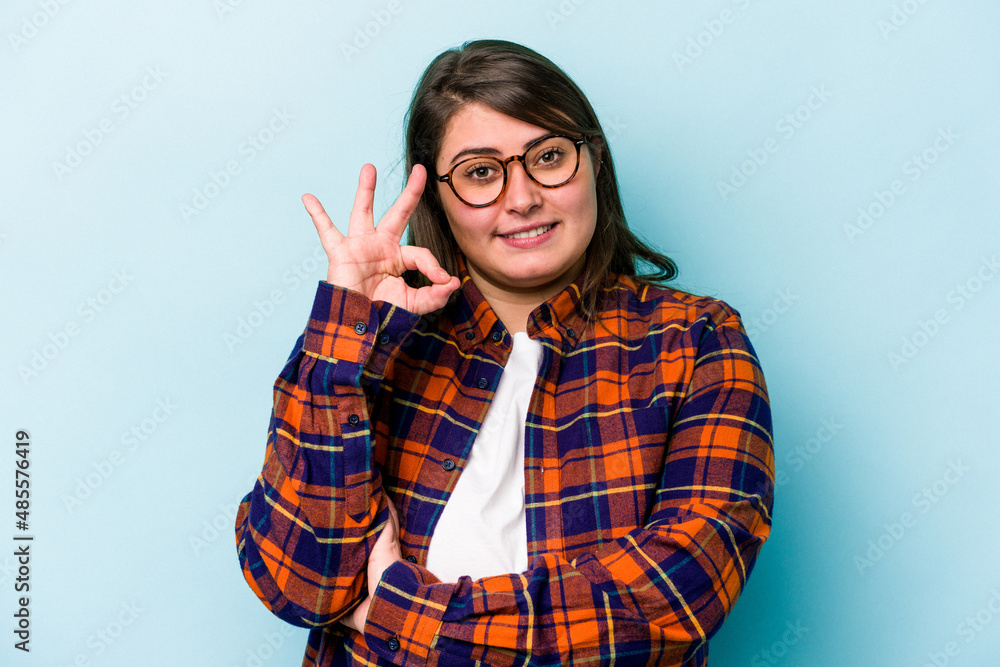 Young caucasian overweight woman isolated on blue background winks an eye and holds an okay gesture with hand. Young caucasian overweight woman isolated on blue background winks an eye and holds an okay gesture with hand.