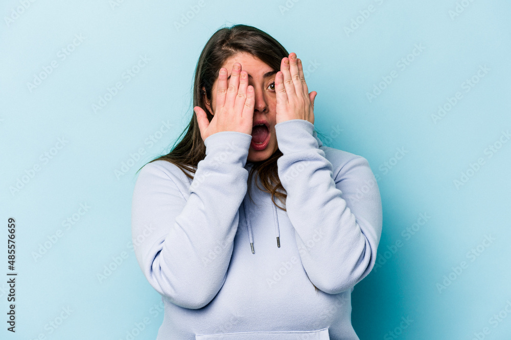 Young caucasian overweight woman isolated on blue background having fun covering half of face with palm. Young caucasian overweight woman isolated on blue background having fun covering half of face with palm.