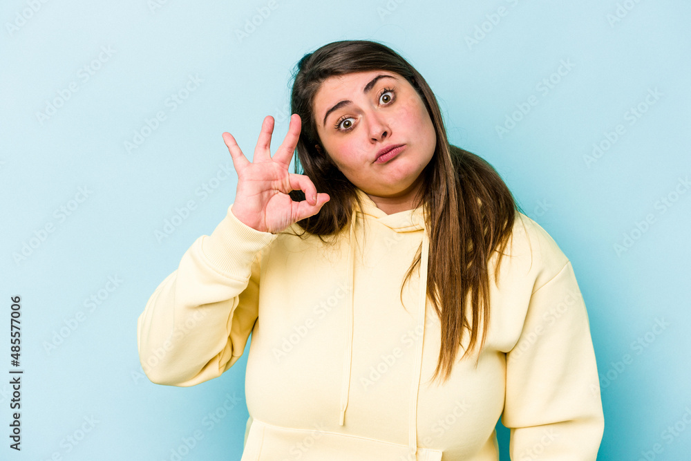 Young caucasian overweight woman isolated on blue background winks an eye and holds an okay gesture with hand. Young caucasian overweight woman isolated on blue background winks an eye and holds an okay gesture with hand.