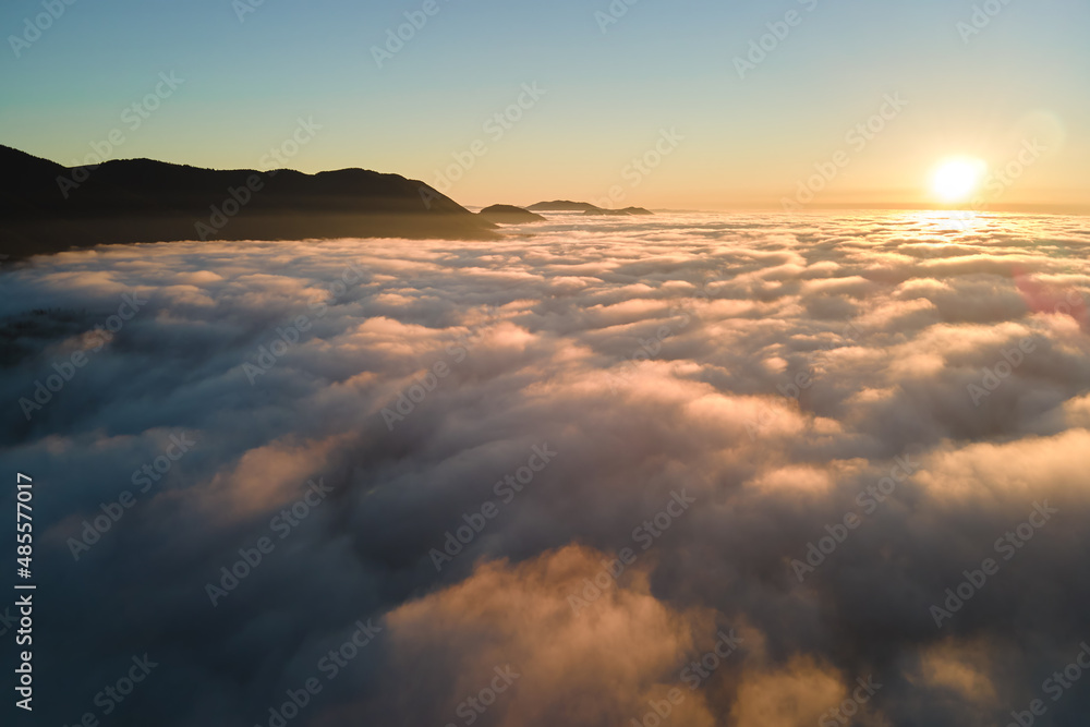 Aerial view of colorful sunrise over white dense fog with distant dark silhouettes of mountain hills on horizon