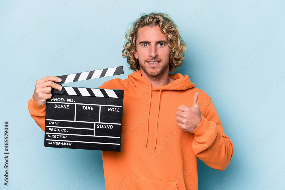 Young caucasian man holding a clapperboard isolated on blue background smiling and raising thumb up