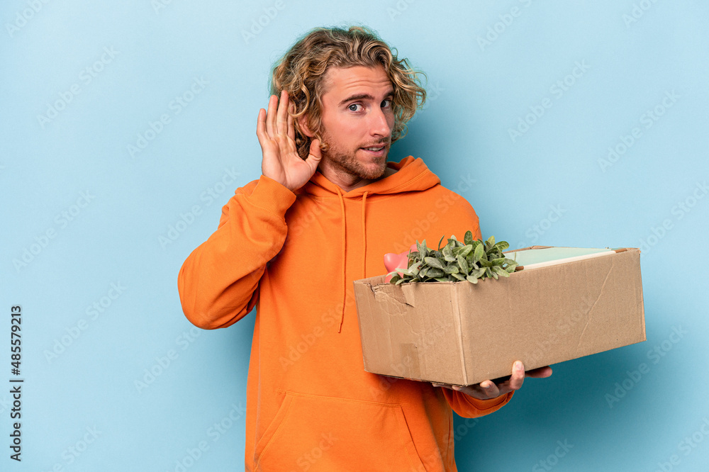 Young caucasian man making a move while picking up a box full of things isolated on blue background trying to listening a gossip. Young caucasian man making a move while picking up a box full of things isolated on blue background trying to listening a gossip.
