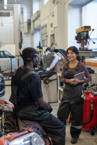 Vertical portrait of two smiling car mechanics chatting in garage shop, focus on young woman in workwear