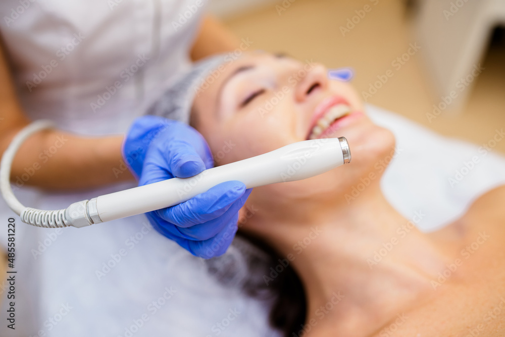 Close-up top-view side-view portrait of a young woman with a towel on her head lying on a table with closed eyes getting a laser skin treatment in healthy beauty spa salon Close-up top-view side-view portrait of a young woman with a towel on her head lying on a table with closed eyes getting a laser skin treatment in healthy beauty spa salon
