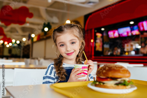 A little curly haired girl sits at the table looking at hamburgers and french fries  thinking about whether to eat. unhealthy food Childhood concepts and eating.