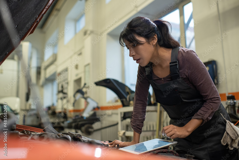 Side view portrait of young female mechanic looking under hood while inspecting vehicle in car repair shop, copy space Side view portrait of young female mechanic looking under hood while inspecting vehicle in car repair shop, copy space