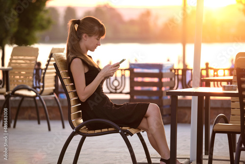 Young woman sitting at street cafe table browsing her cellphone outdoors on warm summer evening. Communication and mobile connection concept.