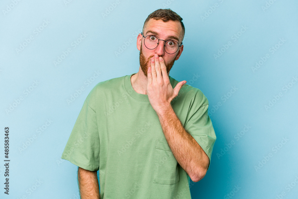 Young caucasian man isolated on blue background shocked, covering mouth with hands, anxious to discover something new. Young caucasian man isolated on blue background shocked, covering mouth with hands, anxious to discover something new.