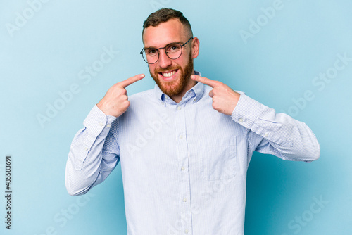 Young caucasian man isolated on blue background smiles, pointing fingers at mouth.