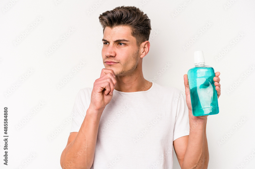 Young caucasian man holding mouthwash isolated on white background looking sideways with doubtful and skeptical expression. Young caucasian man holding mouthwash isolated on white background looking sideways with doubtful and skeptical expression.
