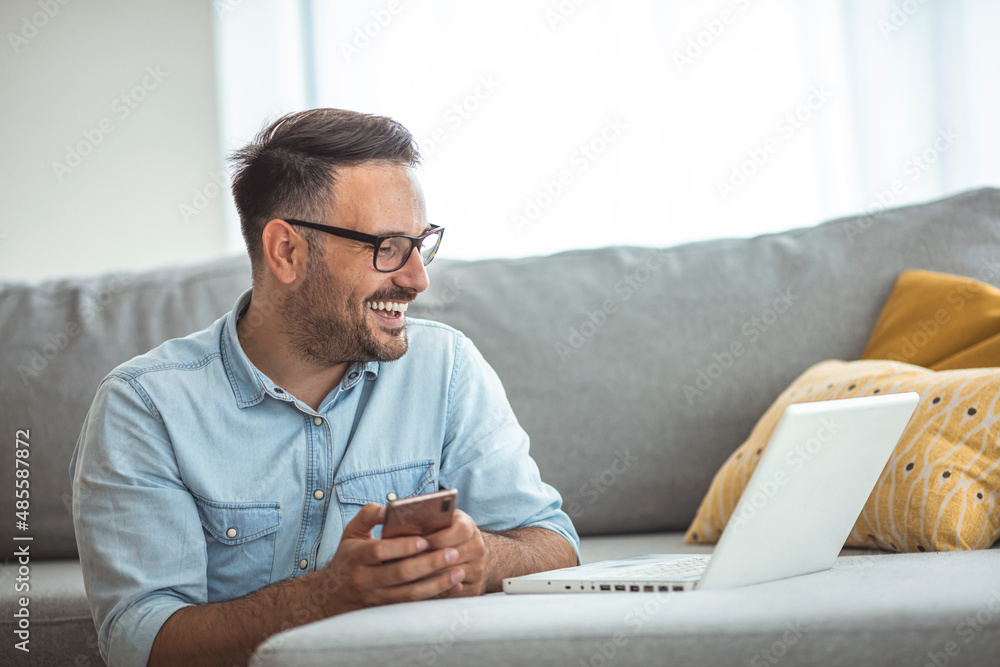 A young attractive smiling guy examines his laptop and holds a mobile phone, sits at home on a comfortable sofa, dressed in casual clothes. The concept of free business work from home A young attractive smiling guy examines his laptop and holds a mobile phone, sits at home on a comfortable sofa, dressed in casual clothes. The concept of free business work from home