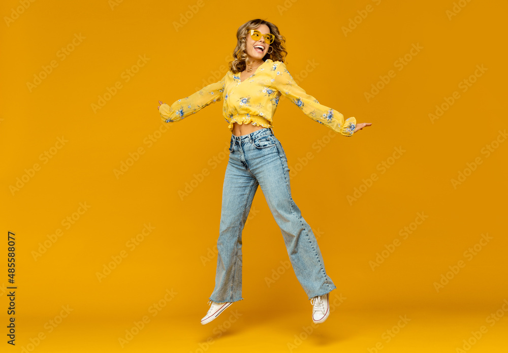 stylish happy smiling blond woman posing in jeans on yellow background