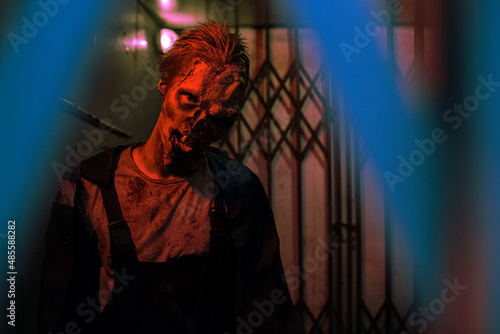 Waist up shot of gory zombie looking at camera in dark hallway lit by red light, copy space