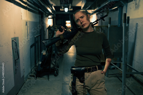 Front view portrait of tough female fighter with big gun looking at camera while standing in dark industrial hallway, copy space