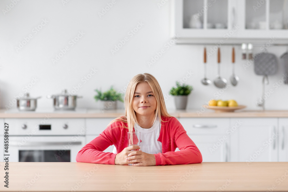 Smiling caucasian teenage girl blonde sits at table with glass of water in modern minimalist kitchen interior Smiling caucasian teenage girl blonde sits at table with glass of water in modern minimalist kitchen interior