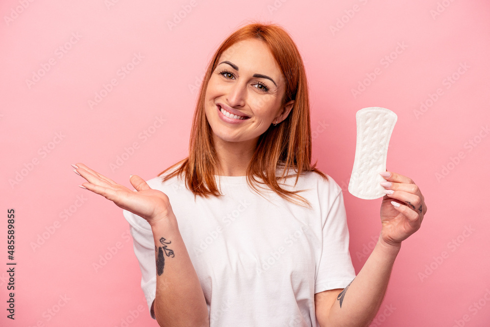 Young caucasian woman holding sanitary napkin isolated on pink background showing a copy space on a palm and holding another hand on waist. Young caucasian woman holding sanitary napkin isolated on pink background showing a copy space on a palm and holding another hand on waist.
