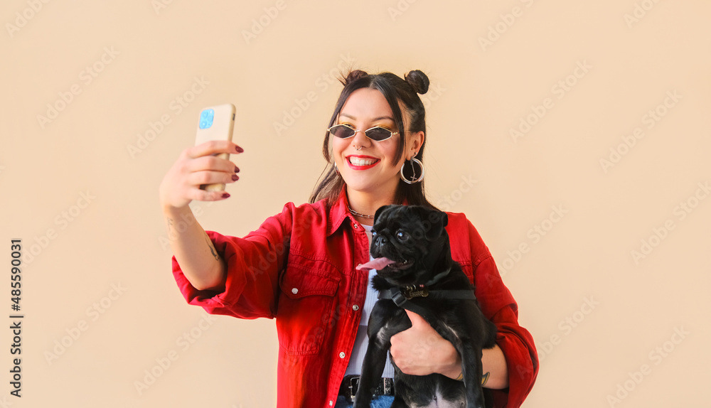Unusual trendy woman with tattoos poses makes a selfie online life video stream or a photo on a mobile phone with her pet dog, a black pug Unusual trendy woman with tattoos poses makes a selfie online life video stream or a photo on a mobile phone with her pet dog, a black pug