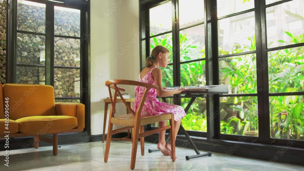 Girl teen playing the piano in a beautiful modern home. Child is learning to play the electronic piano against the background of a glass wall and green garden. Girl teen playing the piano in a beautiful modern home. Child is learning to play the electronic piano against the background of a glass wall and green garden.
