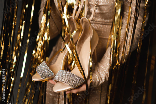 Cropped image of woman holding shoes high heels, prepare for prom ballroom party celebration.Gold lametta on background.