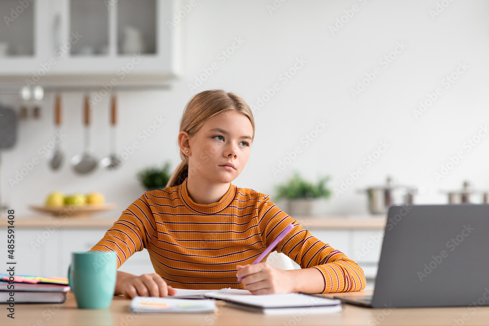 Serious caucasian teenage girl blond sits at table, looks at laptop, studies in minimalist kitchen Serious caucasian teenage girl blond sits at table, looks at laptop, studies in minimalist kitchen