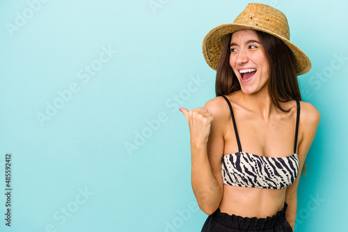 Young caucasian woman going to the beach wearing bikini isolated on blue background points with thumb finger away, laughing and carefree.