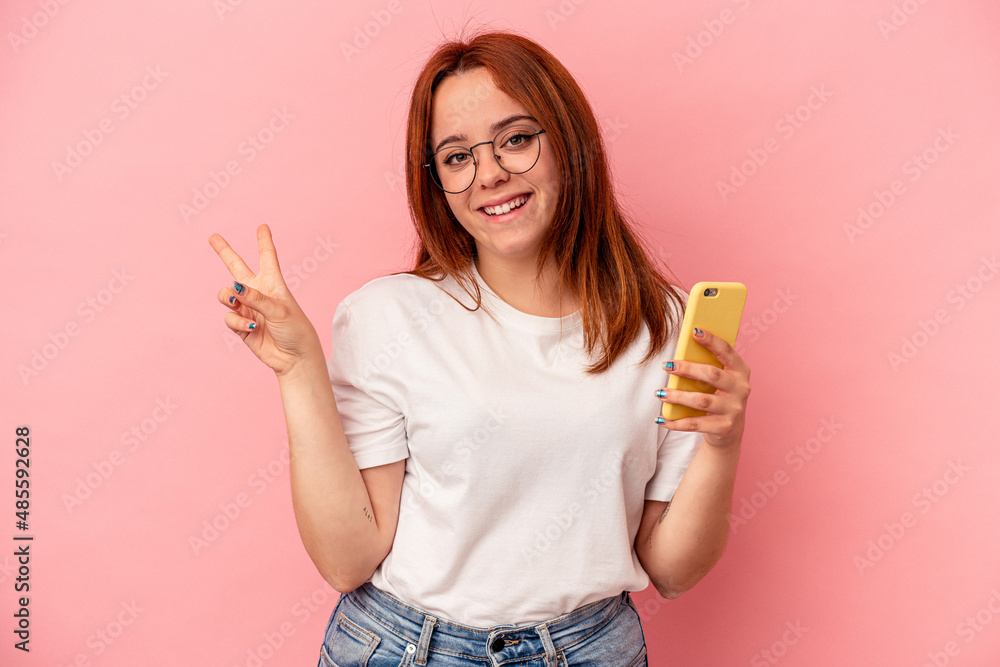 Young caucasian woman holding a mobile phone isolated on pink background joyful and carefree showing a peace symbol with fingers. Young caucasian woman holding a mobile phone isolated on pink background joyful and carefree showing a peace symbol with fingers.