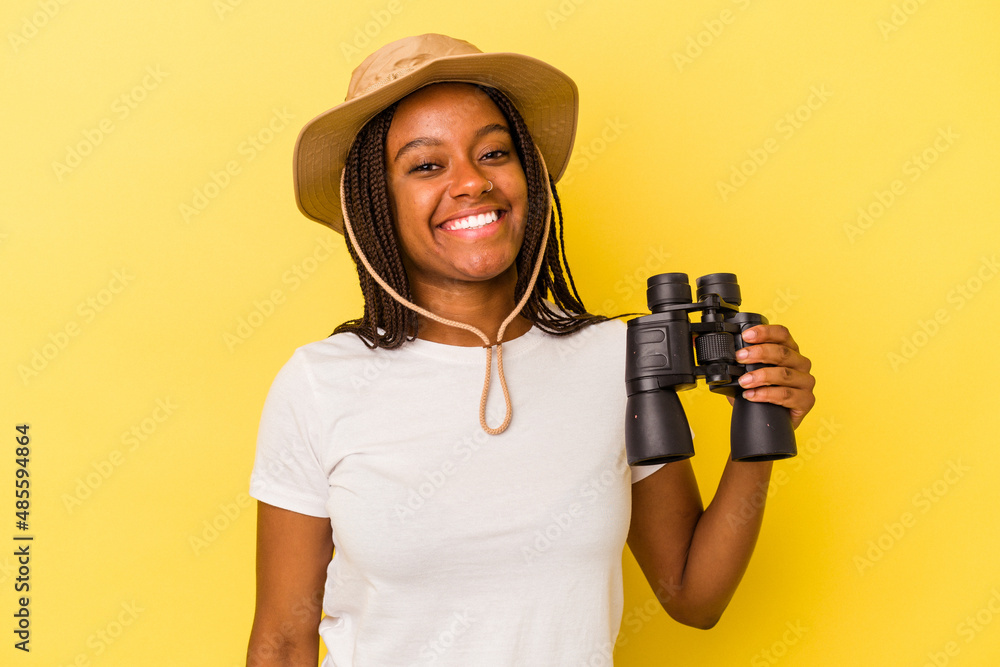 Young african american explorer woman holding a binoculars isolated on yellow background happy, smiling and cheerful. Young african american explorer woman holding a binoculars isolated on yellow background happy, smiling and cheerful.