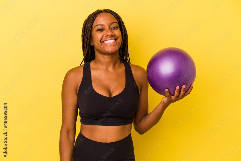 Young african american woman holding a pilates ball isolated on yellow background happy, smiling and cheerful. Young african american woman holding a pilates ball isolated on yellow background happy, smiling and cheerful.