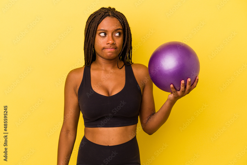Young african american woman holding a pilates ball isolated on yellow background confused, feels doubtful and unsure. Young african american woman holding a pilates ball isolated on yellow background confused, feels doubtful and unsure.
