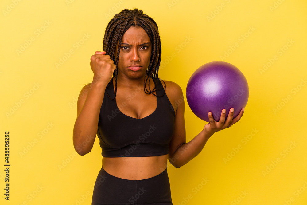 Young african american woman holding a pilates ball isolated on yellow background showing fist to camera, aggressive facial expression. Young african american woman holding a pilates ball isolated on yellow background showing fist to camera, aggressive facial expression.