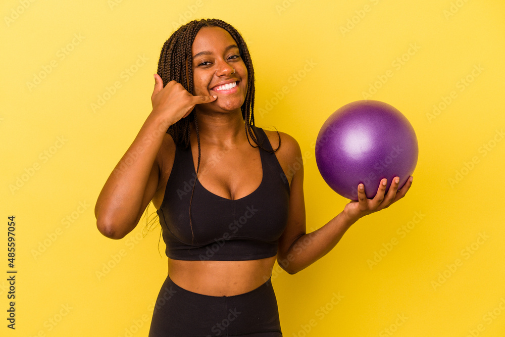 Young african american woman holding a pilates ball isolated on yellow background showing a mobile phone call gesture with fingers. Young african american woman holding a pilates ball isolated on yellow background showing a mobile phone call gesture with fingers.