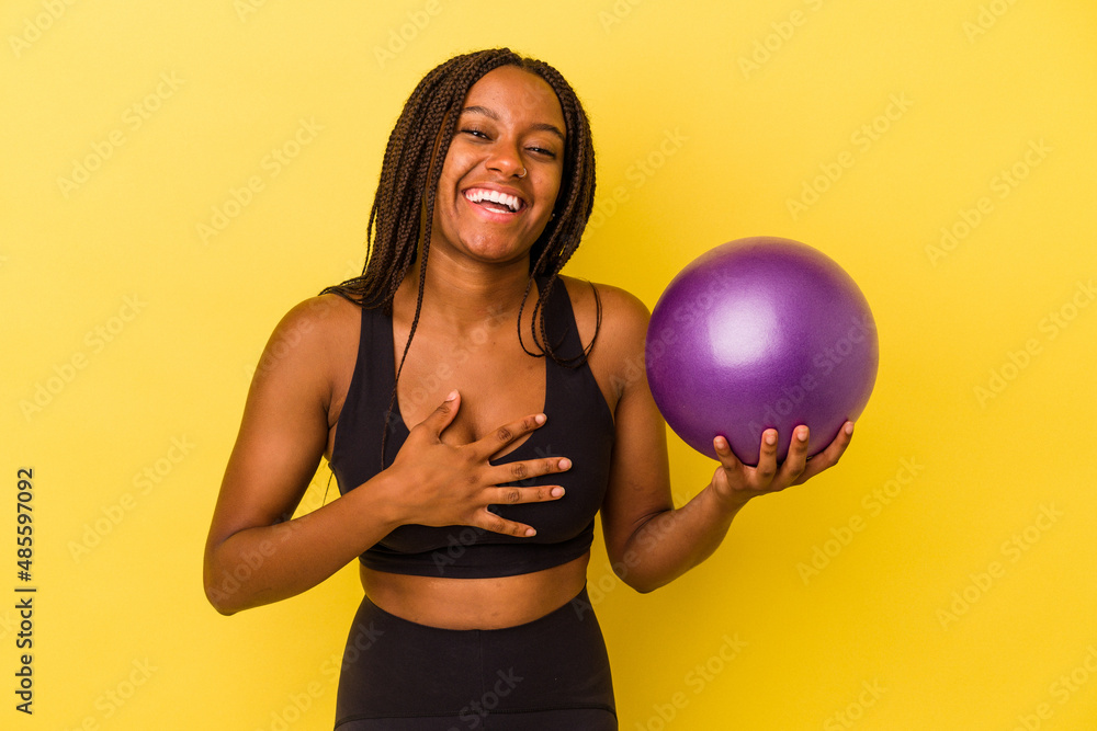 Young african american woman holding a pilates ball isolated on yellow background laughs out loudly keeping hand on chest. Young african american woman holding a pilates ball isolated on yellow background laughs out loudly keeping hand on chest.