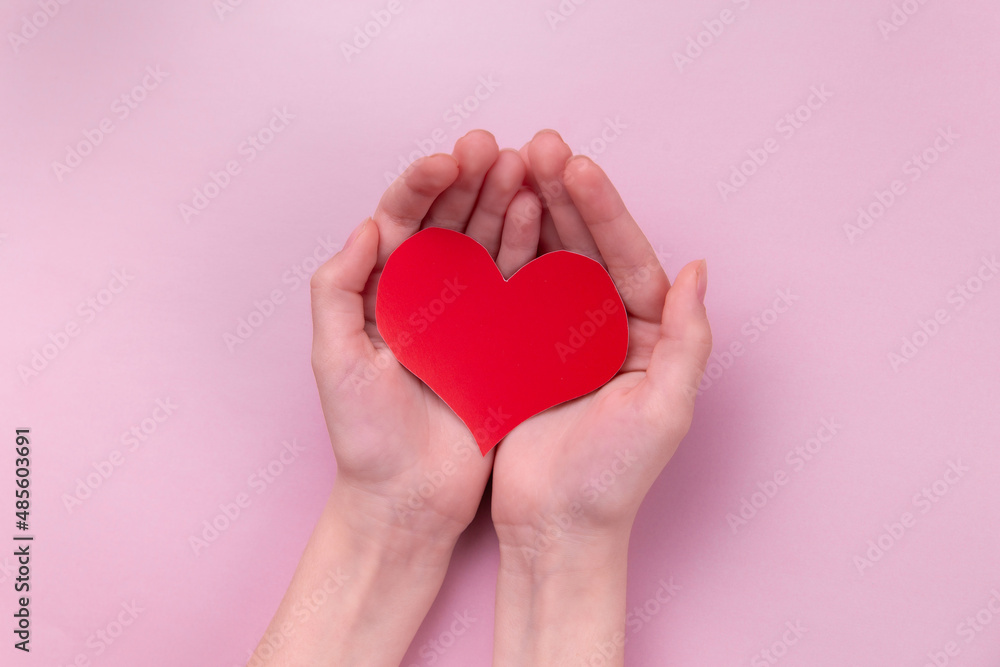 Hands of a young girl holding a red heart on a pink background, top view.