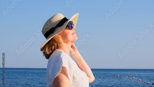 Happy mature woman 50 years old dressed in white dress, straw beach and sunglasses on the beach near the seashore. Vacation concept, relaxation, retirement age