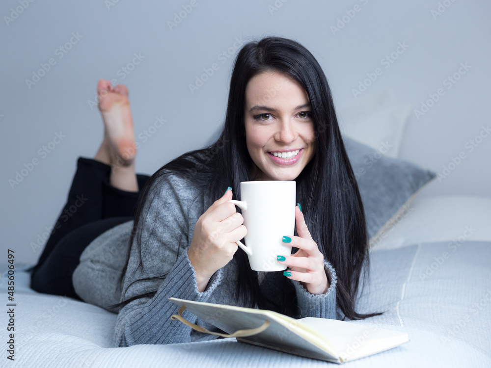 Selective focus horizontal portrait of beautiful smiling brunette young woman in grey sweater and dark pants lying on bed with cup of hot beverage and notebook Selective focus horizontal portrait of beautiful smiling brunette young woman in grey sweater and dark pants lying on bed with cup of hot beverage and notebook