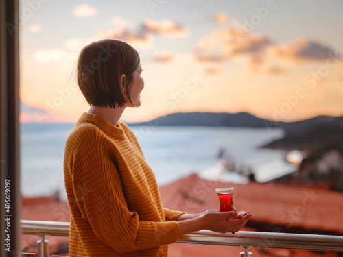 Woman drinking turkish tea from traditional turkish teacup and enjoys panorama over sunset of Kas resort town of Mediterranean sea in Turkey © YURII Seleznov