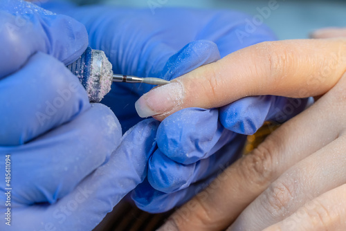Close-up of the hands of a manicure master in blue gloves using an electric nail file to trim and remove cuticles in a beauty salon. Hardware manicure