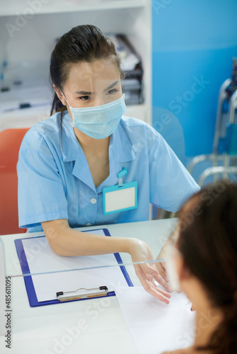 Young medical nurse in protectiove mask explaining patient how to fill form with personal data