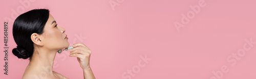 Side view of asian woman massaging neck with jade roller isolated on pink, banner