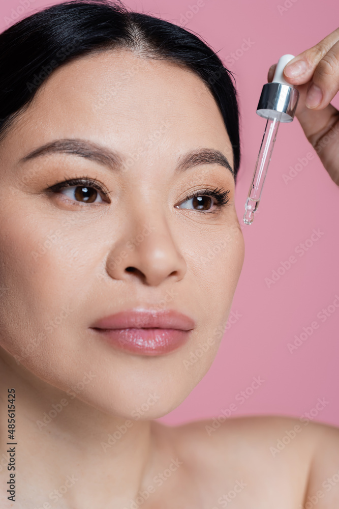 Close up view of asian woman applying serum on face isolated on pink