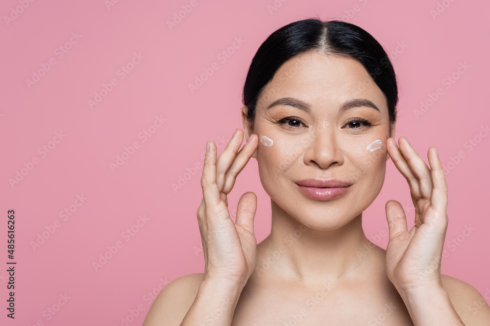 Cheerful asian woman applying face cream on cheeks isolated on pink Cheerful asian woman applying face cream on cheeks isolated on pink