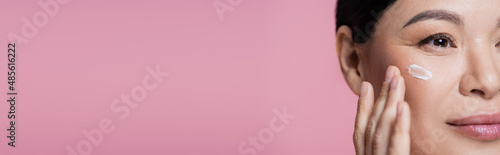 Cropped view of asian woman applying face cream on cheek isolated on pink, banner