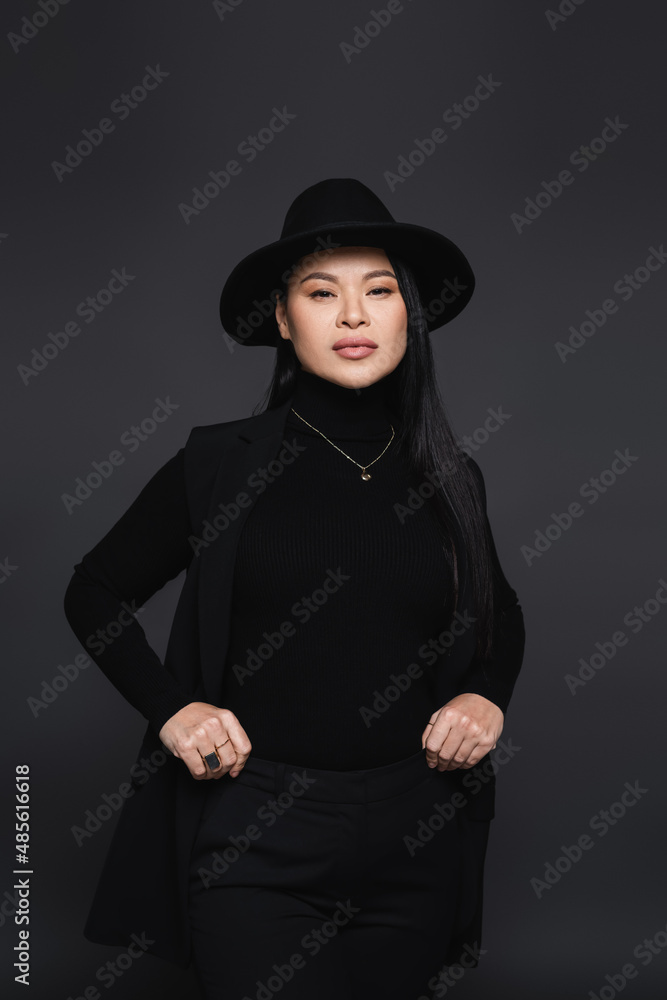 Stylish asian woman in fedora hat and turtleneck looking at camera isolated on dark grey Stylish asian woman in fedora hat and turtleneck looking at camera isolated on dark grey