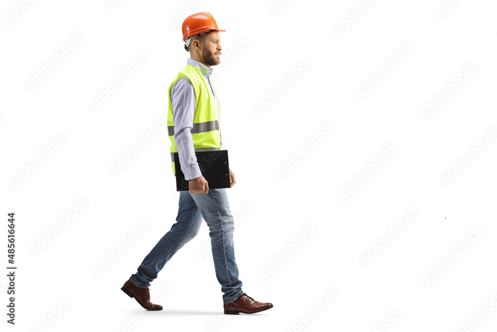Full length profile shot of an engineer with a hardhat walking and holding a clipboard Full length profile shot of an engineer with a hardhat walking and holding a clipboard