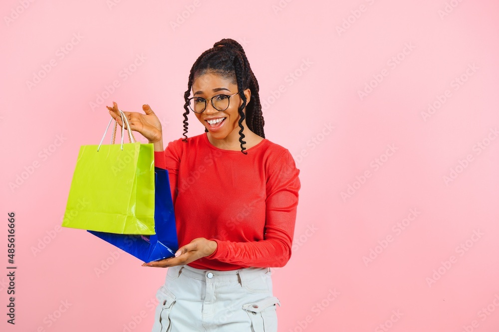 Big Sale. Excited afro girl with shopping bags touching sunglasses, looking at camera over pink studio wall