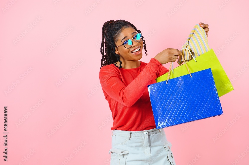 Fototapeta premium Young black woman holding shopping bags on pink backgrond
