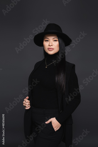 Stylish asian woman in fedora hat posing and looking at camera isolated on dark grey © LIGHTFIELD STUDIOS